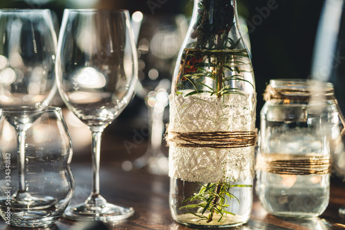 Decorated outdoor restaurant table setting prepared for fine dining with empty wine glasses and glass bottles filled with Rosemarie along with plates, cutlery and other food decor. 