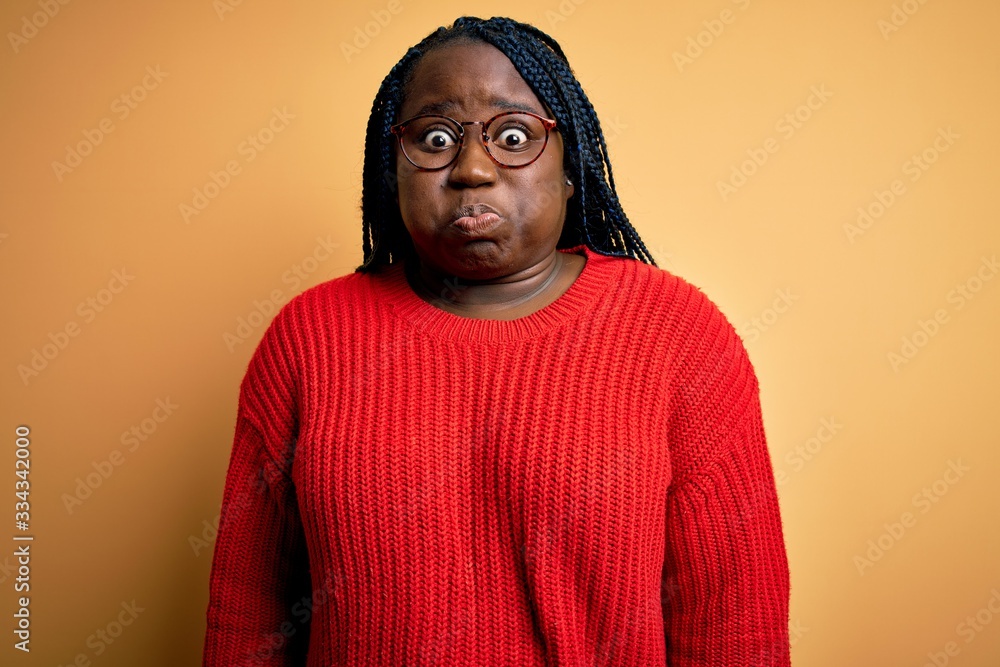 African american plus size woman with braids wearing casual sweater over yellow background puffing cheeks with funny face. Mouth inflated with air, crazy expression. African american plus size woman with braids wearing casual sweater over yellow background puffing cheeks with funny face. Mouth inflated with air, crazy expression.