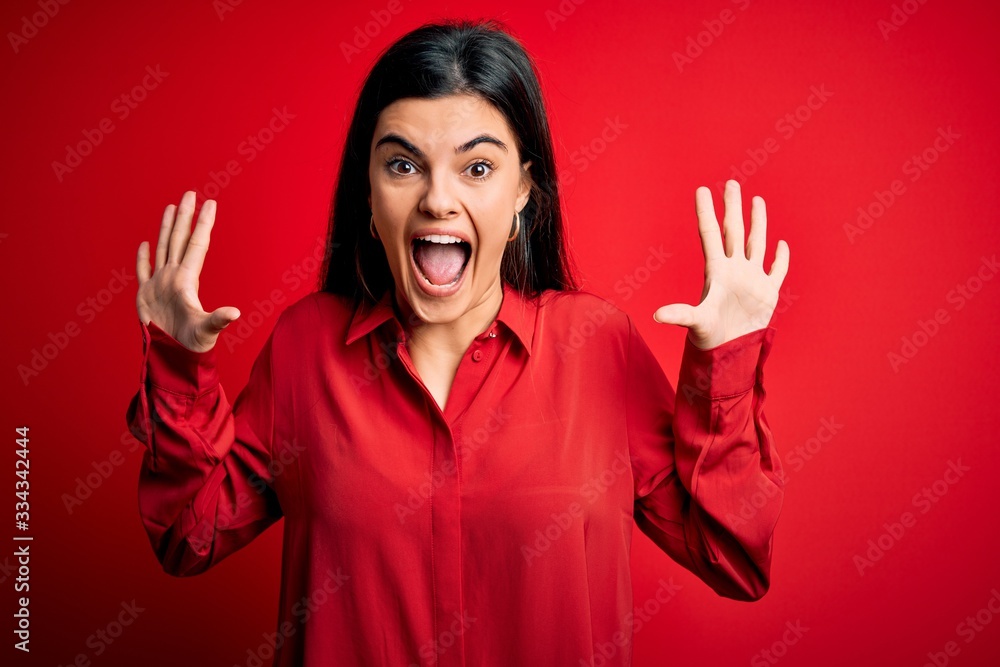 Young beautiful brunette woman wearing casual shirt standing over red background celebrating crazy and amazed for success with arms raised and open eyes screaming excited. Winner concept Young beautiful brunette woman wearing casual shirt standing over red background celebrating crazy and amazed for success with arms raised and open eyes screaming excited. Winner concept
