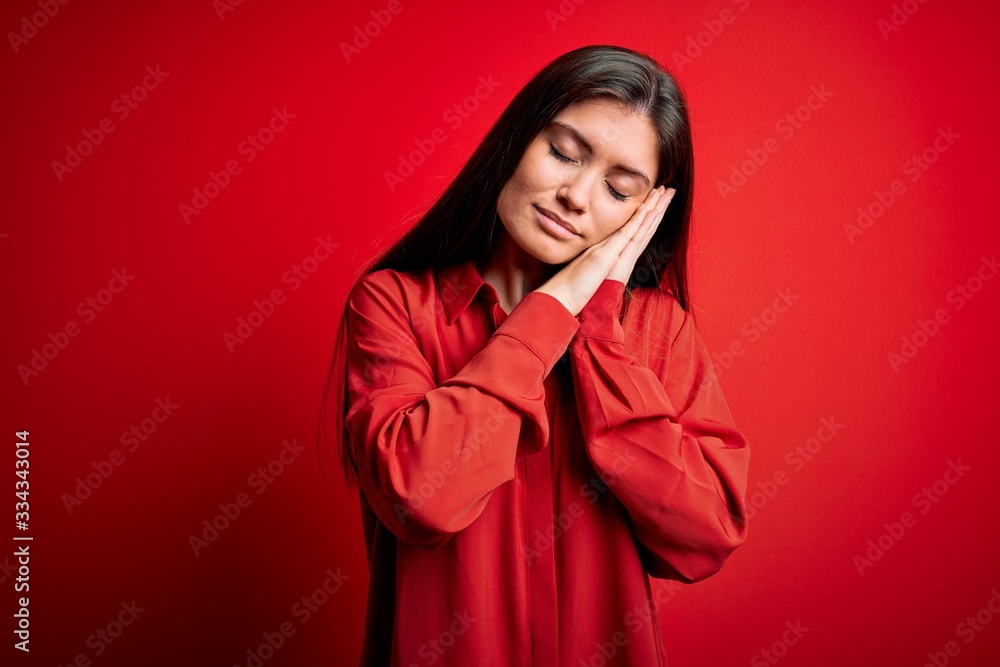 Young beautiful woman with blue eyes wearing casual shirt standing over red background sleeping tired dreaming and posing with hands together while smiling with closed eyes. Young beautiful woman with blue eyes wearing casual shirt standing over red background sleeping tired dreaming and posing with hands together while smiling with closed eyes.