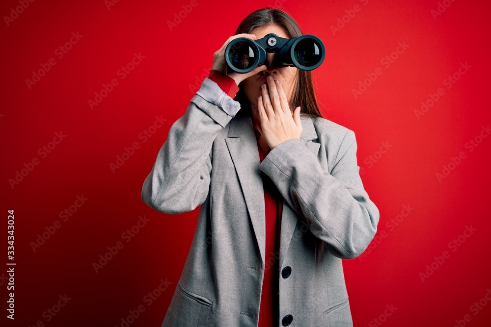Young beautiful woman with blue eyes using binoculars over isolated red background cover mouth with hand shocked with shame for mistake, expression of fear, scared in silence, secret concept Young beautiful woman with blue eyes using binoculars over isolated red background cover mouth with hand shocked with shame for mistake, expression of fear, scared in silence, secret concept