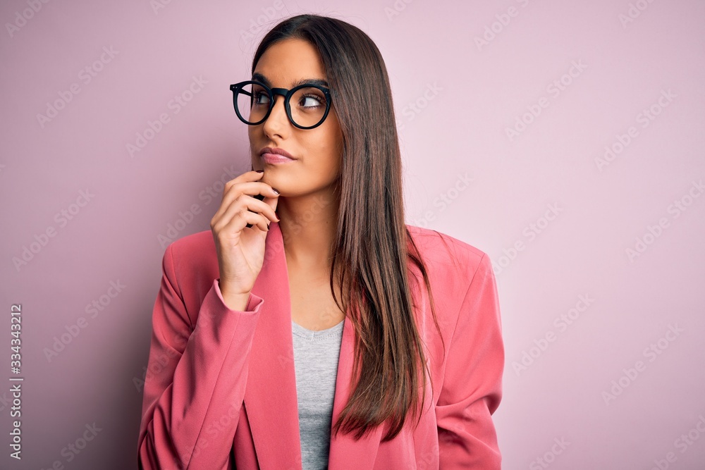 Young beautiful brunette businesswoman wearing jacket and glasses over pink background with hand on chin thinking about question, pensive expression. Smiling with thoughtful face. Doubt concept. Young beautiful brunette businesswoman wearing jacket and glasses over pink background with hand on chin thinking about question, pensive expression. Smiling with thoughtful face. Doubt concept.