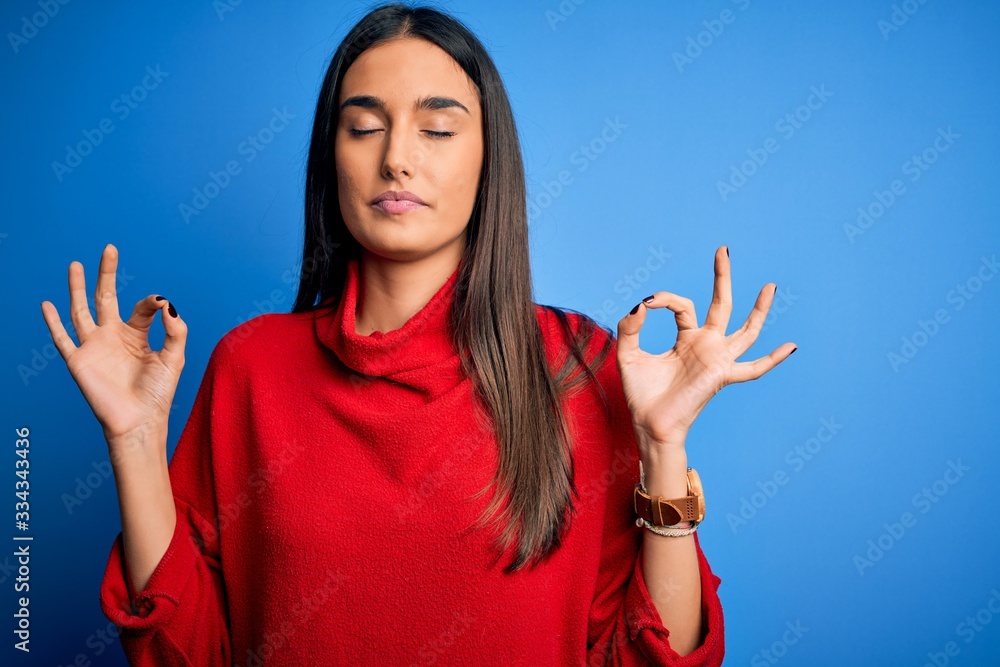 Young beautiful brunette woman wearing red casual sweater over isolated blue background relax and smiling with eyes closed doing meditation gesture with fingers. Yoga concept. Young beautiful brunette woman wearing red casual sweater over isolated blue background relax and smiling with eyes closed doing meditation gesture with fingers. Yoga concept.