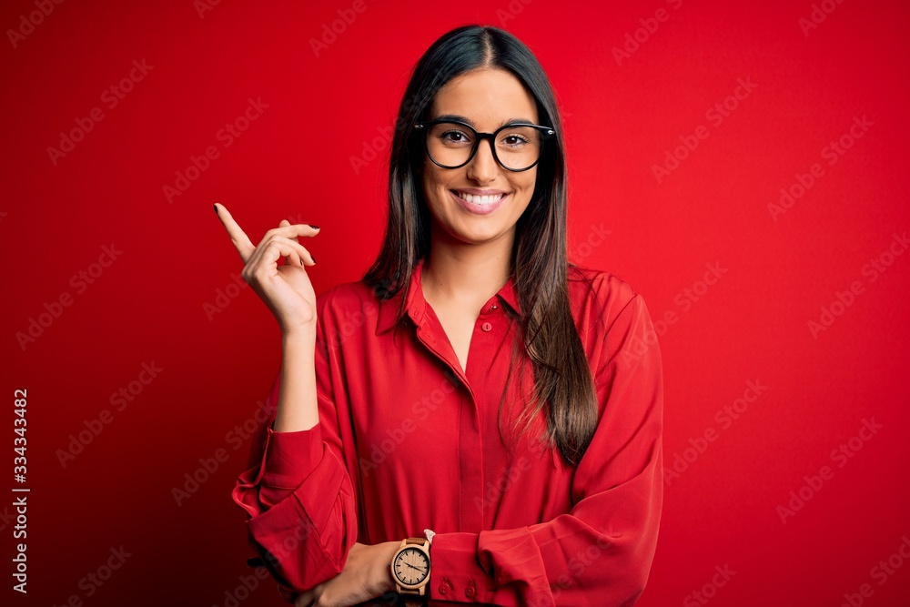 Φωτογραφία Young beautiful brunette woman wearing casual shirt and glasses over red background with a big smile on face, pointing with hand and finger to the side looking at the camera