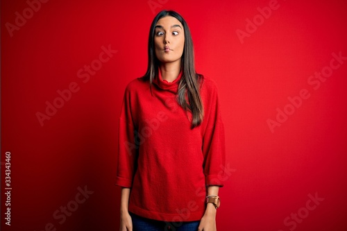 Young beautiful brunette woman wearing casual sweater over isolated red background making fish face with lips, crazy and comical gesture. Funny expression. © Krakenimages.com