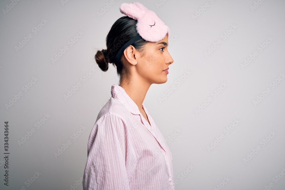 Young beautiful brunette woman wearing pajama and sleep mask over white background looking to side, relax profile pose with natural face with confident smile. Young beautiful brunette woman wearing pajama and sleep mask over white background looking to side, relax profile pose with natural face with confident smile.