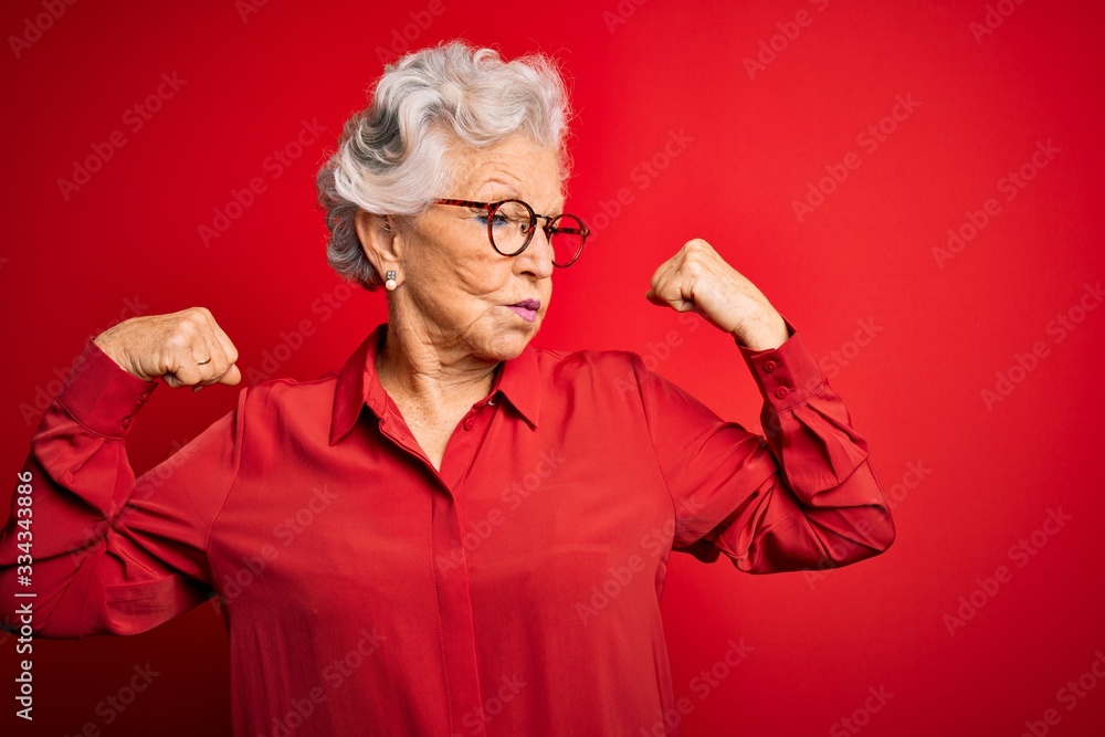 Φωτογραφία Senior beautiful grey-haired woman wearing casual shirt and glasses over red background showing arms muscles smiling proud