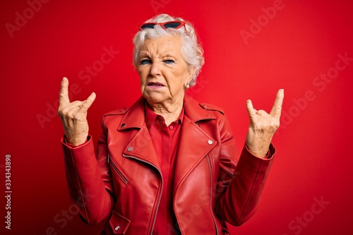 Senior beautiful grey-haired woman wearing casual red jacket and sunglasses shouting with crazy expression doing rock symbol with hands up. Music star. Heavy concept.
