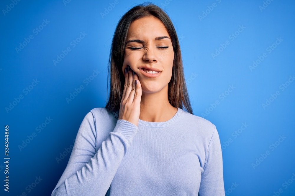 Obraz premium Young beautiful brunette woman wearing casual sweater standing over blue background touching mouth with hand with painful expression because of toothache or dental illness on teeth. Dentist