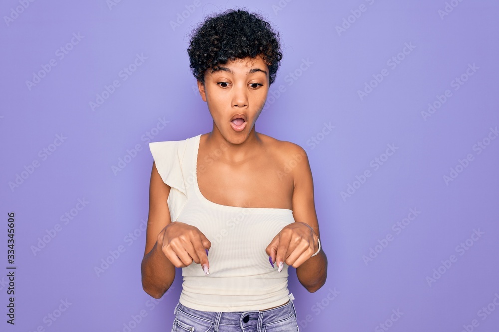 Young beautiful african american afro woman wearing casual t-shirt over purple background Pointing down with fingers showing advertisement, surprised face and open mouth Young beautiful african american afro woman wearing casual t-shirt over purple background Pointing down with fingers showing advertisement, surprised face and open mouth