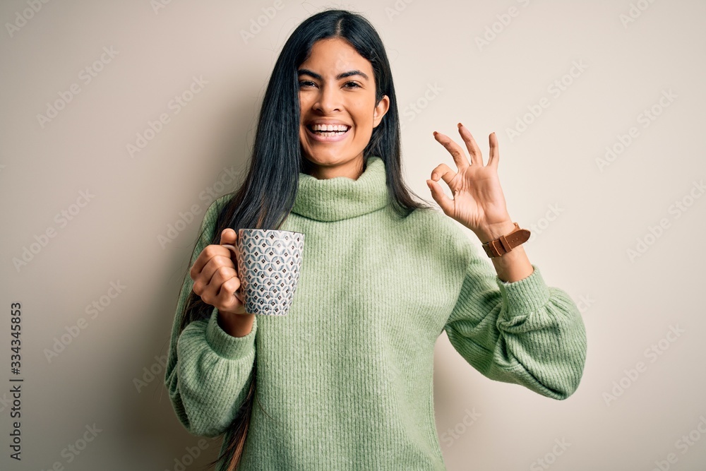 Young beautiful hispanic woman drinking a cup of hot coffee over isolated background doing ok sign with fingers, excellent symbol Young beautiful hispanic woman drinking a cup of hot coffee over isolated background doing ok sign with fingers, excellent symbol