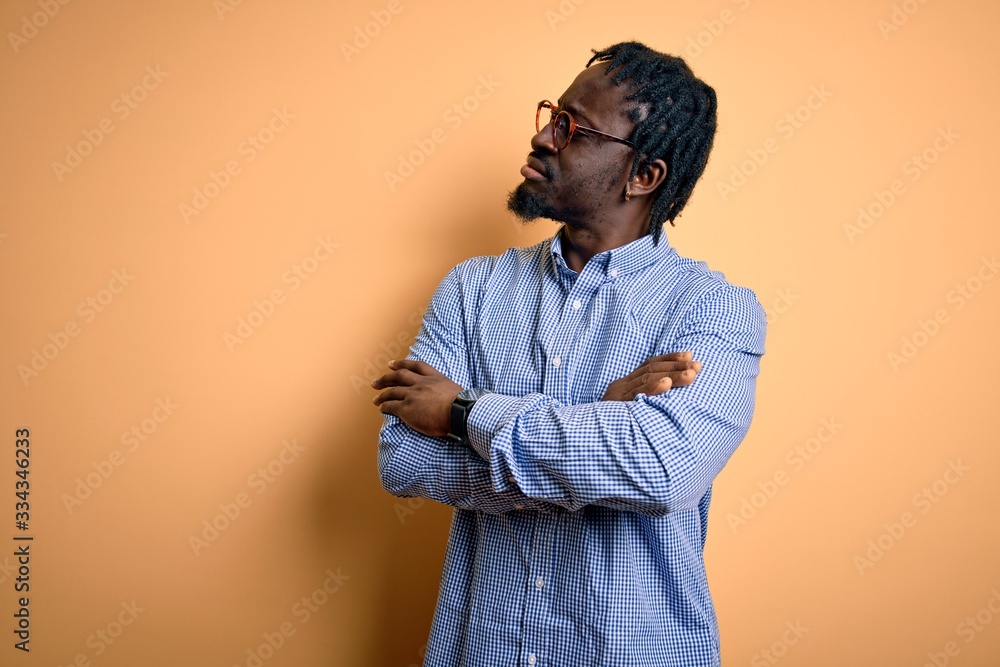 Young handsome african american man wearing shirt and glasses over yellow background looking to the side with arms crossed convinced and confident