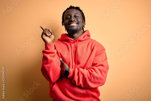 Young african american sporty man wearing sweatshirt with hoodie over yellow background with a big smile on face, pointing with hand and finger to the side looking at the camera.
