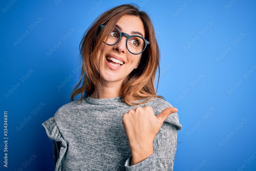 Young beautiful brunette woman wearing casual sweater and glasses over blue background smiling with happy face looking and pointing to the side with thumb up.