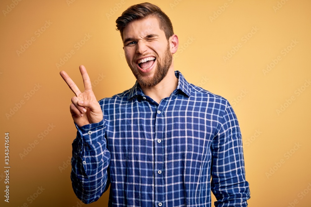 Young blond businessman with beard and blue eyes wearing shirt over yellow background smiling with happy face winking at the camera doing victory sign with fingers. Number two. Young blond businessman with beard and blue eyes wearing shirt over yellow background smiling with happy face winking at the camera doing victory sign with fingers. Number two.