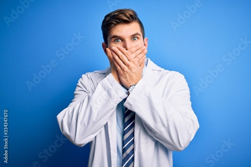 Young blond therapist man with beard and blue eyes wearing coat and tie over background shocked covering mouth with hands for mistake. Secret concept.