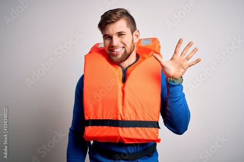Young blond tourist man with beard and blue eyes wearing lifejacket over white background showing and pointing up with fingers number five while smiling confident and happy. © Krakenimages.com