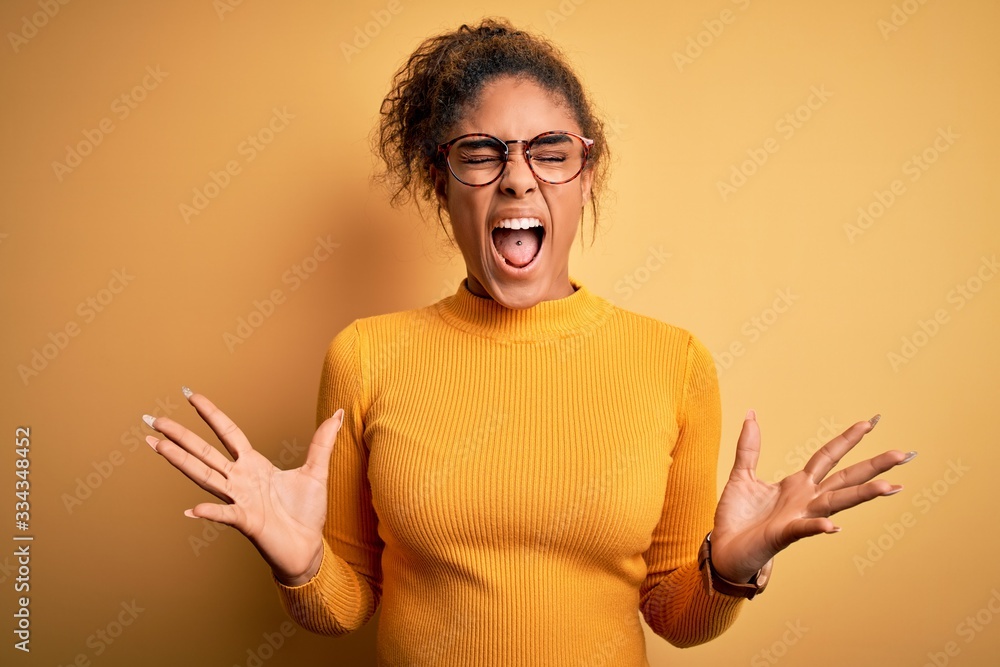 Young beautiful african american girl wearing sweater and glasses over yellow background crazy and mad shouting and yelling with aggressive expression and arms raised. Frustration concept. Young beautiful african american girl wearing sweater and glasses over yellow background crazy and mad shouting and yelling with aggressive expression and arms raised. Frustration concept.