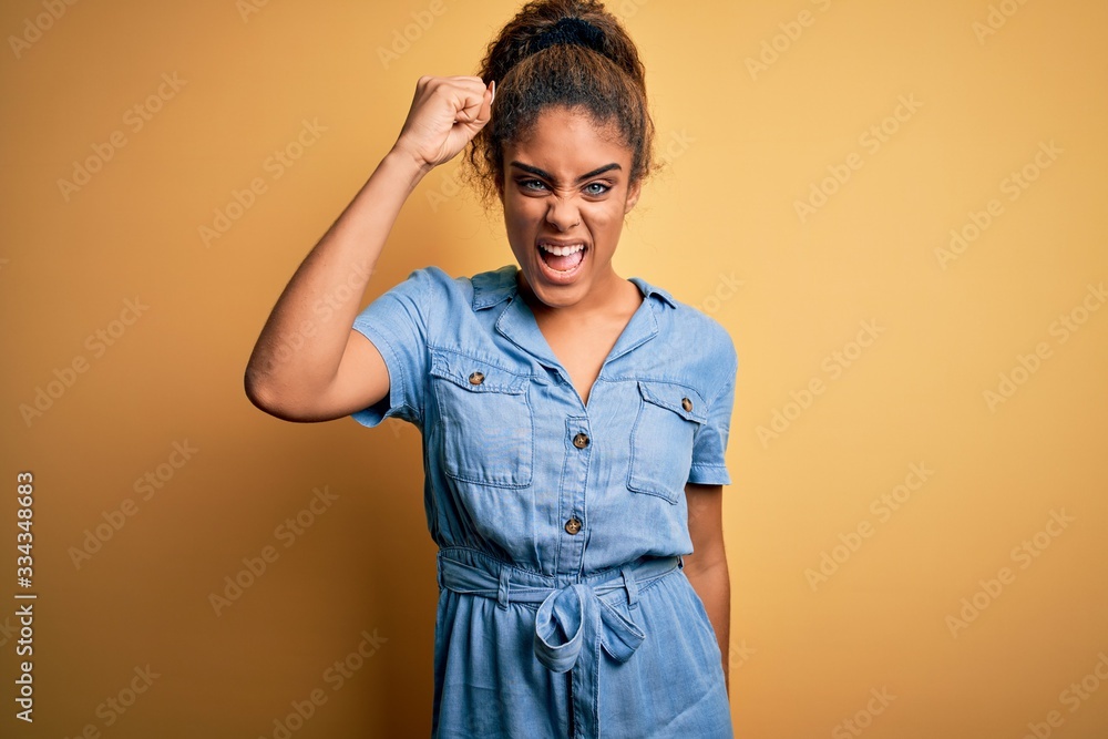 Young beautiful african american girl wearing denim dress standing over yellow background angry and mad raising fist frustrated and furious while shouting with anger. Rage and aggressive concept. Young beautiful african american girl wearing denim dress standing over yellow background angry and mad raising fist frustrated and furious while shouting with anger. Rage and aggressive concept.