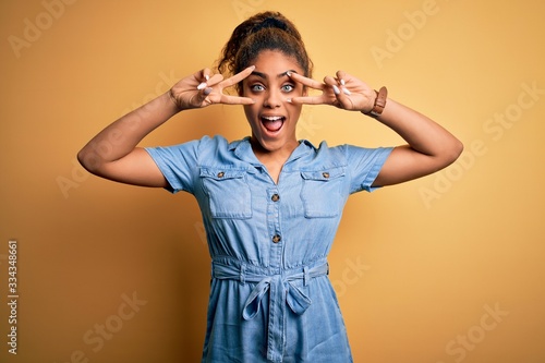 Young beautiful african american girl wearing denim dress standing over yellow background Doing peace symbol with fingers over face, smiling cheerful showing victory