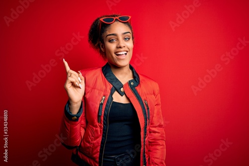 Beautiful african american girl wearing red jacket and sunglasses over isolated background with a big smile on face, pointing with hand and finger to the side looking at the camera.