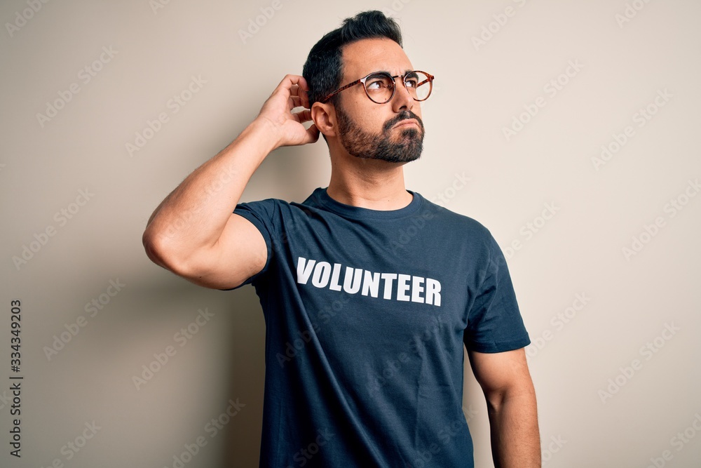 Handsome man with beard wearing t-shirt with volunteer message over white background confuse and wondering about question. Uncertain with doubt, thinking with hand on head. Pensive concept. Handsome man with beard wearing t-shirt with volunteer message over white background confuse and wondering about question. Uncertain with doubt, thinking with hand on head. Pensive concept.