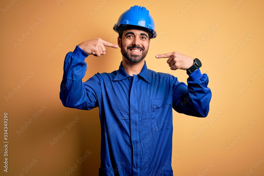 Mechanic man with beard wearing blue uniform and safety helmet over yellow background smiling cheerful showing and pointing with fingers teeth and mouth. Dental health concept.