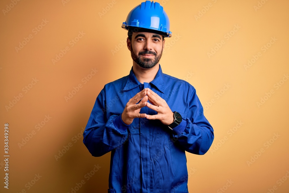 Mechanic man with beard wearing blue uniform and safety helmet over yellow background Hands together and fingers crossed smiling relaxed and cheerful. Success and optimistic