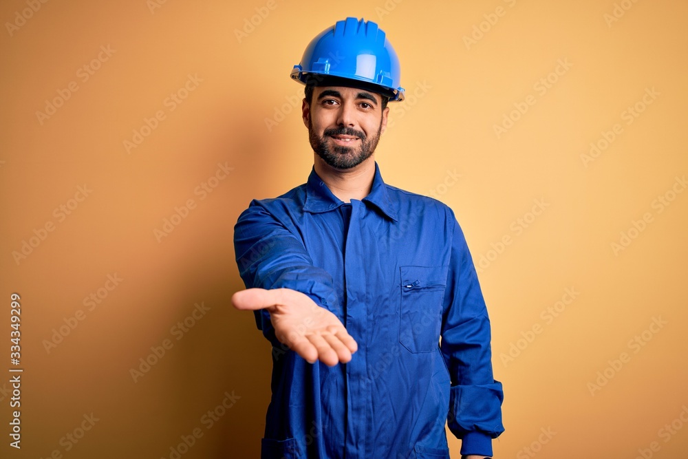 Mechanic man with beard wearing blue uniform and safety helmet over yellow background smiling cheerful offering palm hand giving assistance and acceptance.