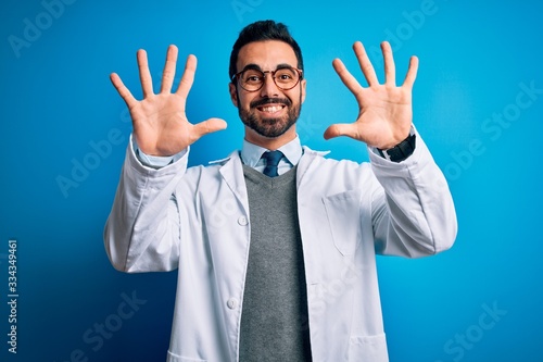 Young handsome doctor man with beard wearing coat and glasses over blue background showing and pointing up with fingers number ten while smiling confident and happy.