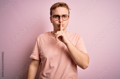Young handsome redhead man wearing casual t-shirt standing over isolated pink background asking to be quiet with finger on lips. Silence and secret concept.