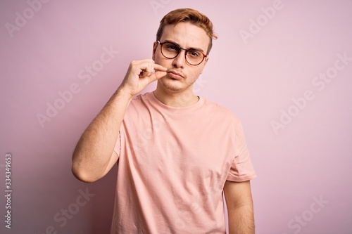 Young handsome redhead man wearing casual t-shirt standing over isolated pink background mouth and lips shut as zip with fingers. Secret and silent, taboo talking © Krakenimages.com