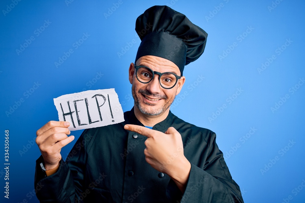 Young handsome chef man overworked wearing uniform and hat holding help paper message very happy pointing with hand and finger Young handsome chef man overworked wearing uniform and hat holding help paper message very happy pointing with hand and finger