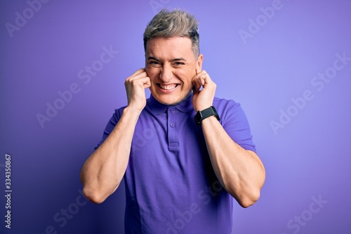 Young handsome modern man wearing casual purple t-shirt over isolated background covering ears with fingers with annoyed expression for the noise of loud music. Deaf concept. © Krakenimages.com