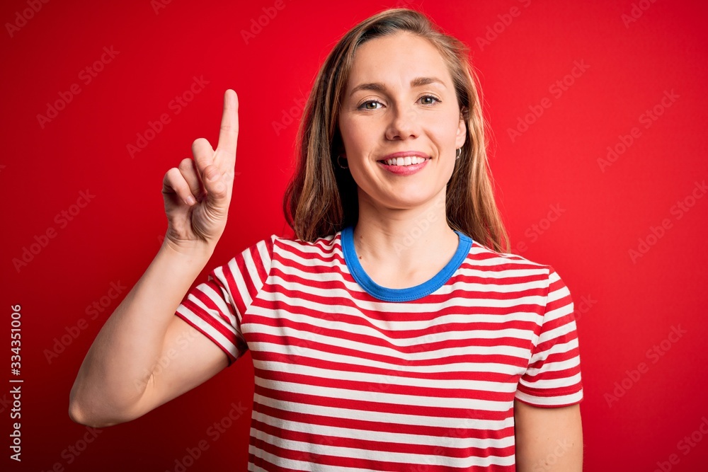 Young beautiful blonde woman wearing casual striped t-shirt over isolated red background showing and pointing up with finger number one while smiling confident and happy. Young beautiful blonde woman wearing casual striped t-shirt over isolated red background showing and pointing up with finger number one while smiling confident and happy.