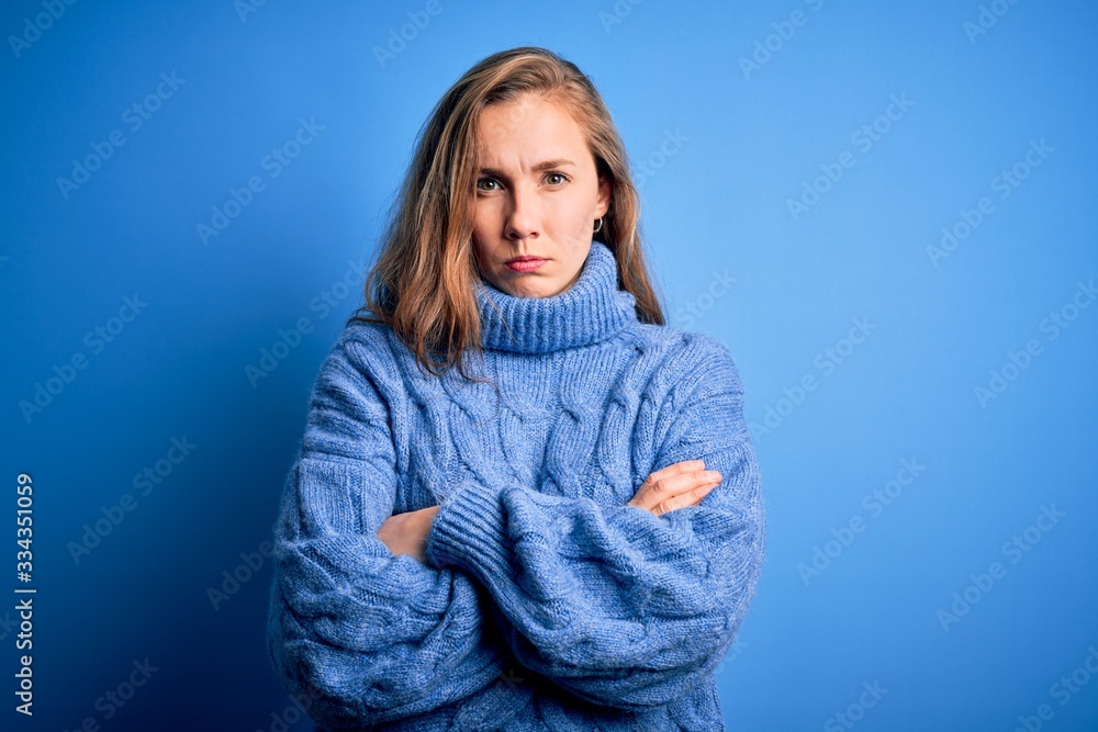 Young beautiful blonde woman wearing casual turtleneck sweater over blue background skeptic and nervous, disapproving expression on face with crossed arms. Negative person. Young beautiful blonde woman wearing casual turtleneck sweater over blue background skeptic and nervous, disapproving expression on face with crossed arms. Negative person.