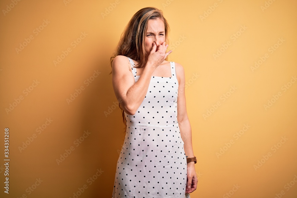 Young beautiful blonde woman on vacation wearing summer dress over yellow background smelling something stinky and disgusting, intolerable smell, holding breath with fingers on nose. Bad smell Young beautiful blonde woman on vacation wearing summer dress over yellow background smelling something stinky and disgusting, intolerable smell, holding breath with fingers on nose. Bad smell