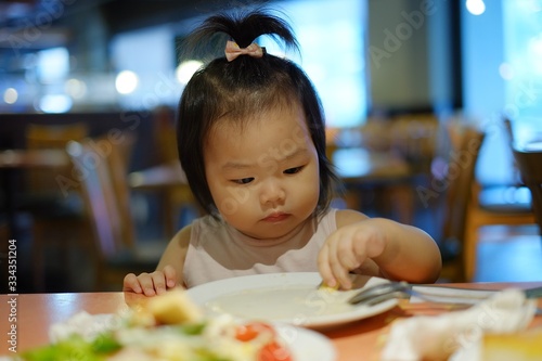 A cute Asian girl eating salad at a restaurant with her mom feeding her using fork and spoon.