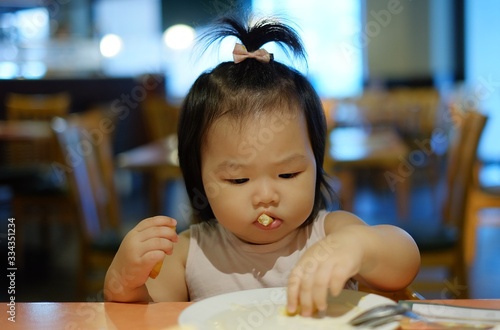 A cute Asian girl eating salad at a restaurant with her mom feeding her using fork and spoon. © Sirichai