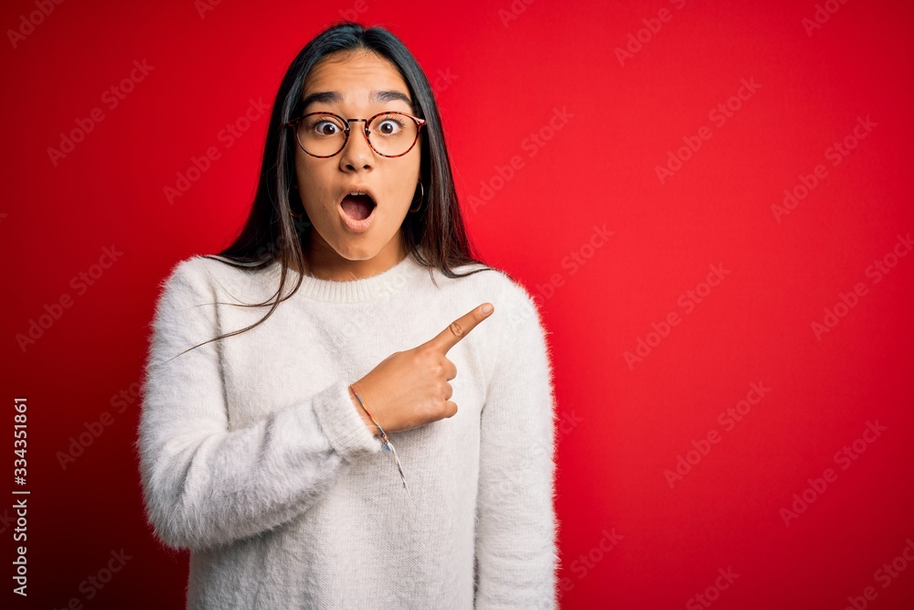 Young beautiful asian woman wearing casual sweater and glasses over red background Surprised pointing with finger to the side, open mouth amazed expression. Young beautiful asian woman wearing casual sweater and glasses over red background Surprised pointing with finger to the side, open mouth amazed expression.