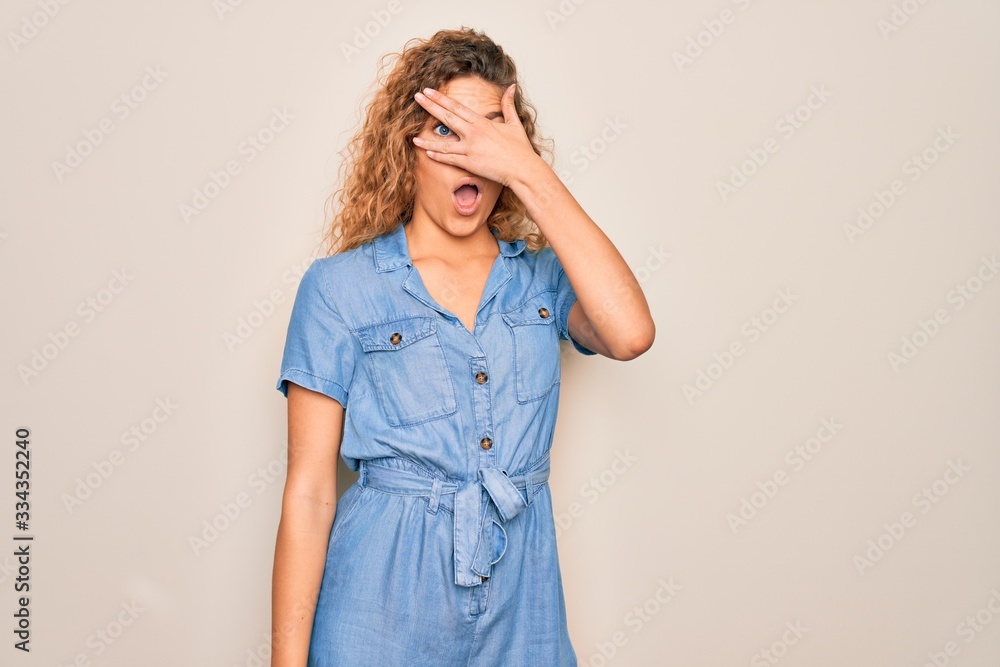 Young beautiful woman with blue eyes wearing casual denim dress over white background peeking in shock covering face and eyes with hand, looking through fingers with embarrassed expression. Young beautiful woman with blue eyes wearing casual denim dress over white background peeking in shock covering face and eyes with hand, looking through fingers with embarrassed expression.