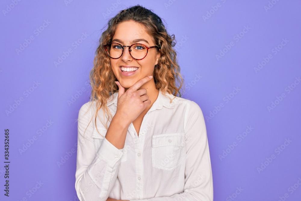 Young beautiful woman with blue eyes wearing casual shirt and glasses over purple background looking confident at the camera smiling with crossed arms and hand raised on chin. Thinking positive.