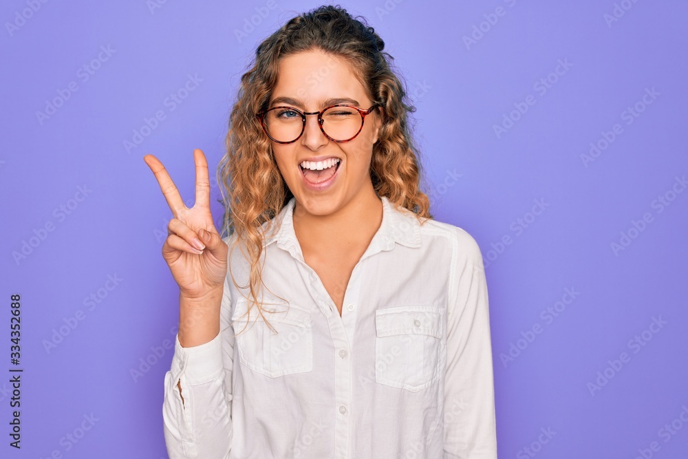Young beautiful woman with blue eyes wearing casual shirt and glasses over purple background smiling with happy face winking at the camera doing victory sign. Number two. Young beautiful woman with blue eyes wearing casual shirt and glasses over purple background smiling with happy face winking at the camera doing victory sign. Number two.