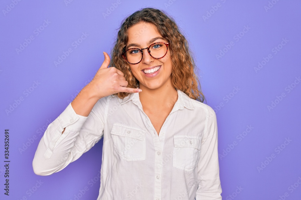 Young beautiful woman with blue eyes wearing casual shirt and glasses over purple background smiling doing phone gesture with hand and fingers like talking on the telephone. Communicating concepts. Young beautiful woman with blue eyes wearing casual shirt and glasses over purple background smiling doing phone gesture with hand and fingers like talking on the telephone. Communicating concepts.