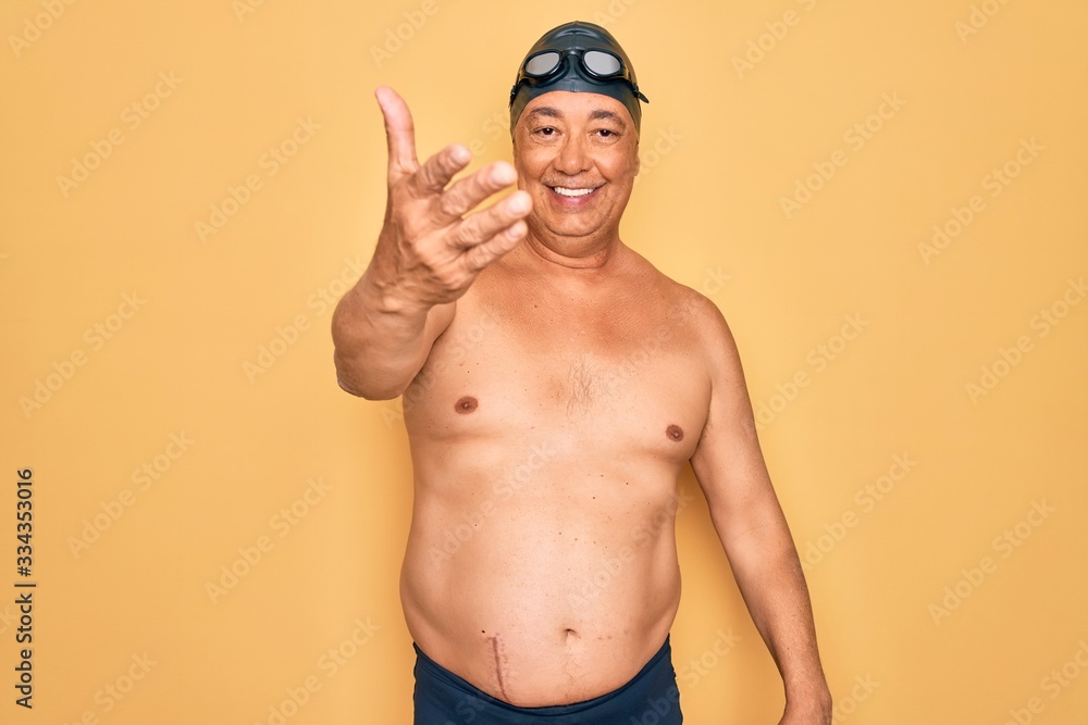 Middle age senior grey-haired swimmer man wearing swimsuit, cap and goggles smiling friendly offering handshake as greeting and welcoming. Successful business.