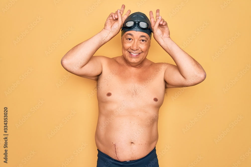 Middle age senior grey-haired swimmer man wearing swimsuit, cap and goggles Posing funny and crazy with fingers on head as bunny ears, smiling cheerful
