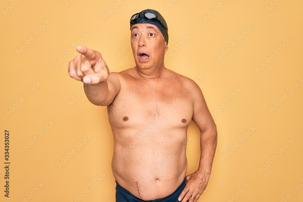 Middle age senior grey-haired swimmer man wearing swimsuit, cap and goggles Pointing with finger surprised ahead, open mouth amazed expression, something on the front