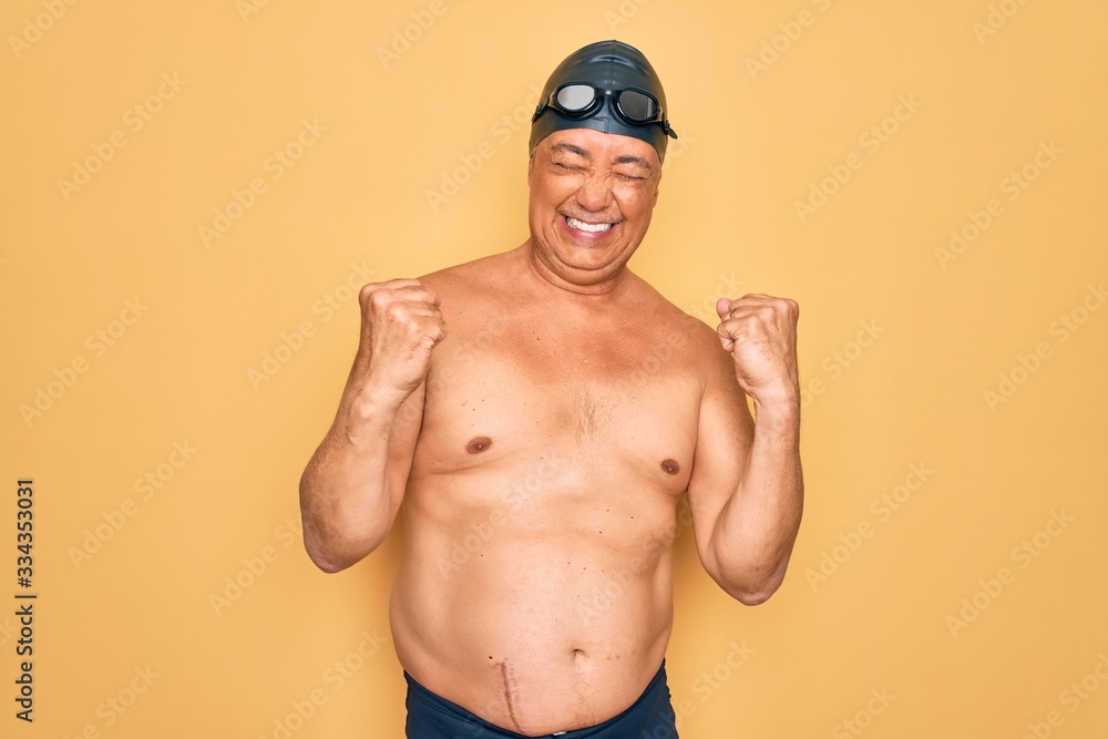 Middle age senior grey-haired swimmer man wearing swimsuit, cap and goggles very happy and excited doing winner gesture with arms raised, smiling and screaming for success. Celebration concept.