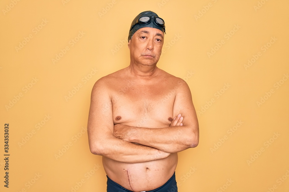 Middle age senior grey-haired swimmer man wearing swimsuit, cap and goggles skeptic and nervous, disapproving expression on face with crossed arms. Negative person.