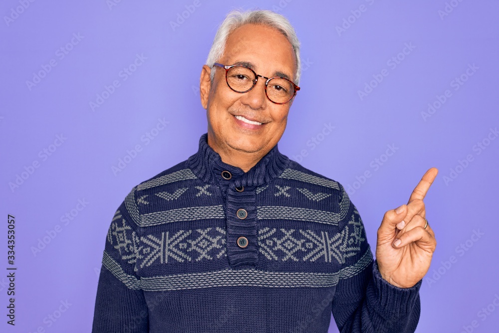 Middle age senior grey-haired man wearing glasses and winter sweater over purple background with a big smile on face, pointing with hand and finger to the side looking at the camera. Middle age senior grey-haired man wearing glasses and winter sweater over purple background with a big smile on face, pointing with hand and finger to the side looking at the camera.